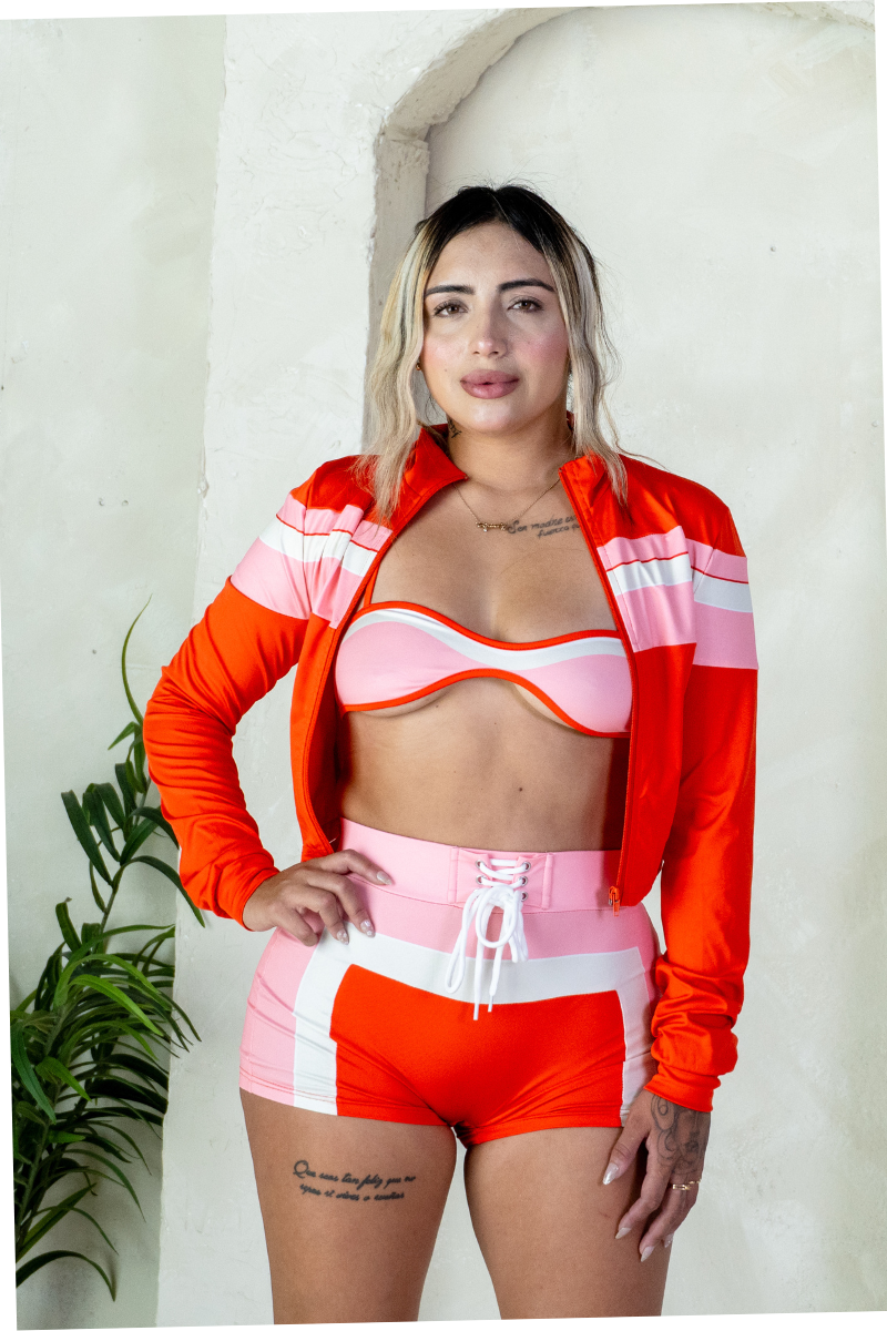 Woman wearing a colorful sports outfit with an orange jacket and pink shorts against a white wall.