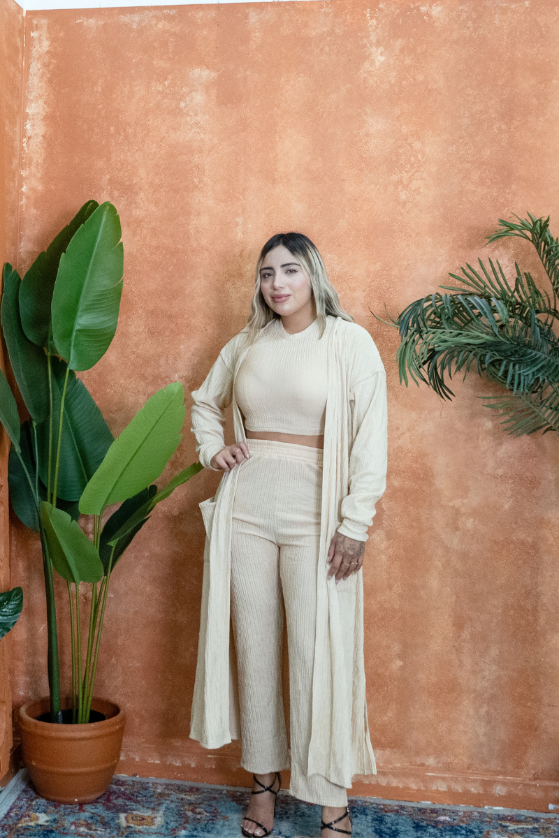 Woman in a beige outfit standing against a textured orange wall with plants.