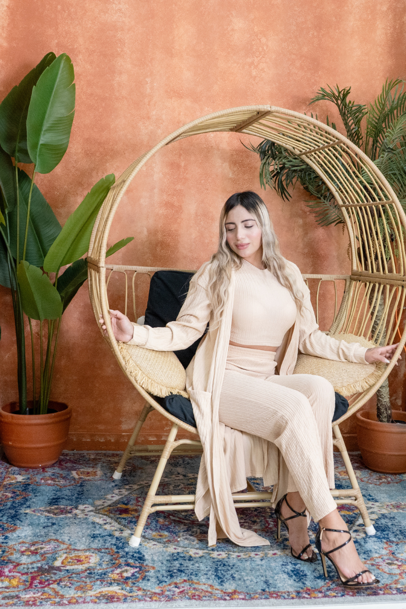 Woman sitting in a circular wicker chair with plants and a colorful rug in the background