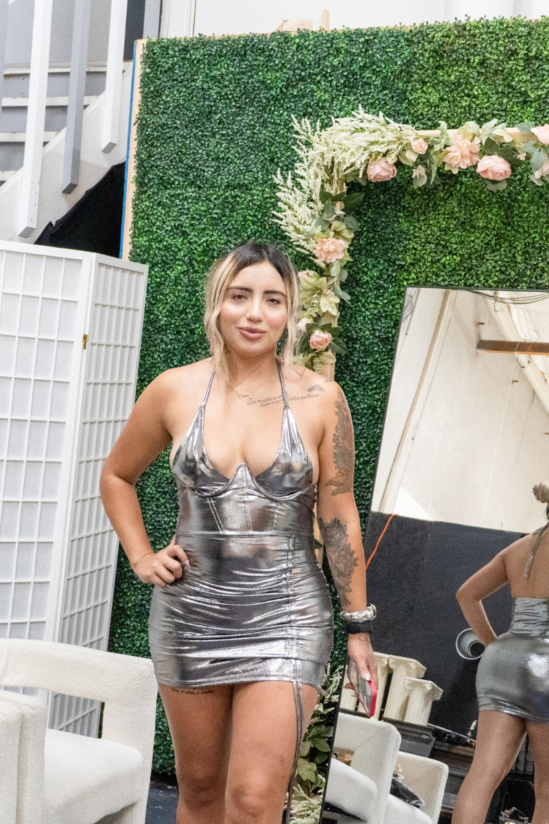 "Woman in a silver dress standing in front of a green wall with floral decorations."