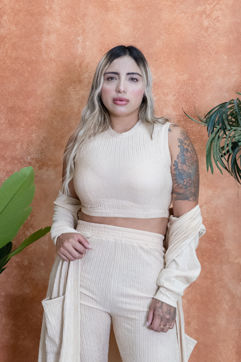 Woman in a cream-colored outfit standing against a textured orange wall with plants.